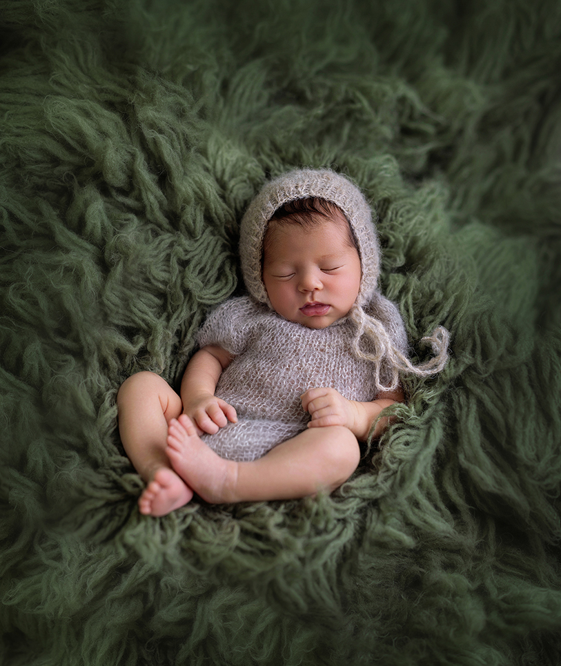 Sleeping newborn baby girl wearing a gray knit bonnet and outfit, peacefully posed on a deep green textured backdrop during a studio newborn photography session.