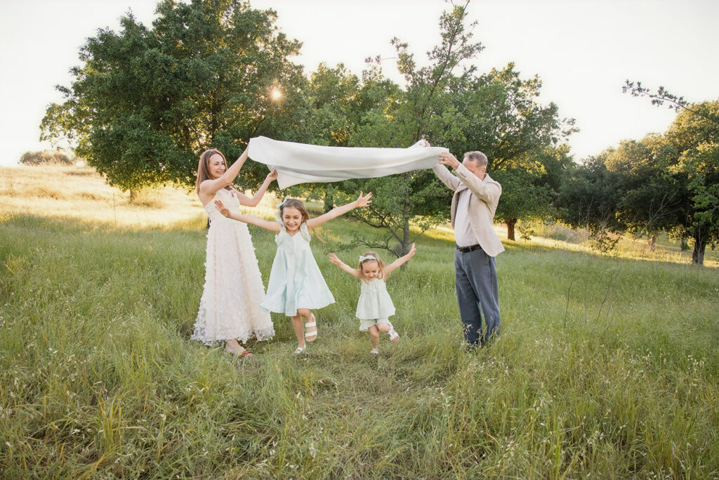 Parents and two daughters holding a flowing white blanket together in a sunlit meadow during an outdoor family photography session.