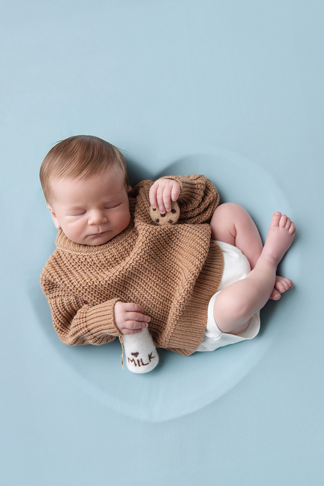 Newborn baby in a warm brown knit outfit peacefully posed beside a small teddy bear on a soft blue background.