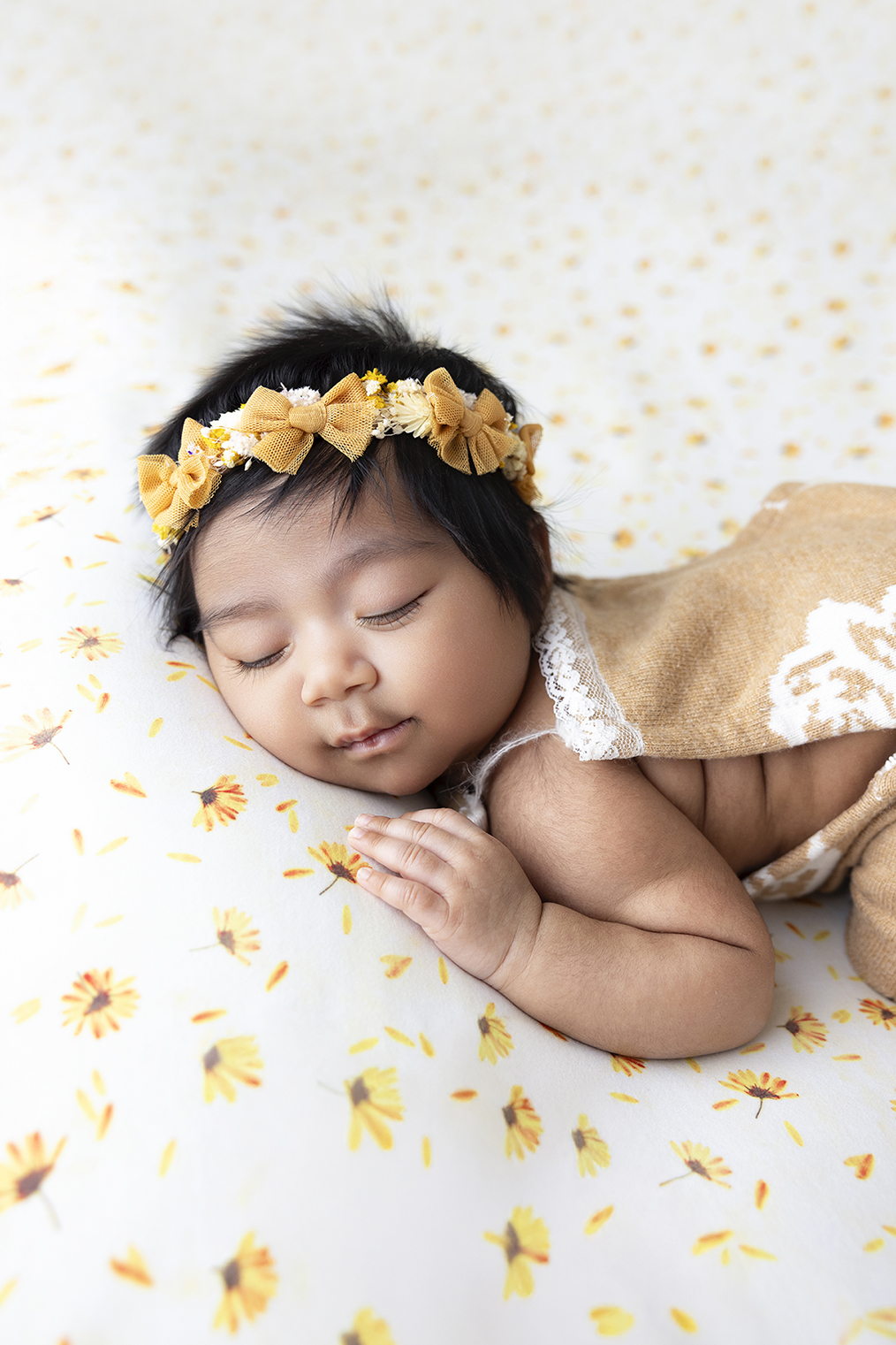 Sleeping newborn baby girl wearing a delicate floral headband and brown outfit, peacefully posed on a soft patterned backdrop.