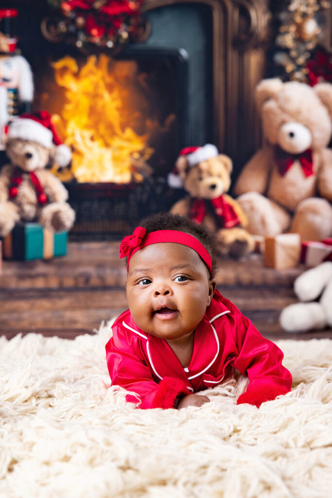 Baby wearing a red Christmas outfit and headband in front of a cozy holiday fireplace with teddy bears