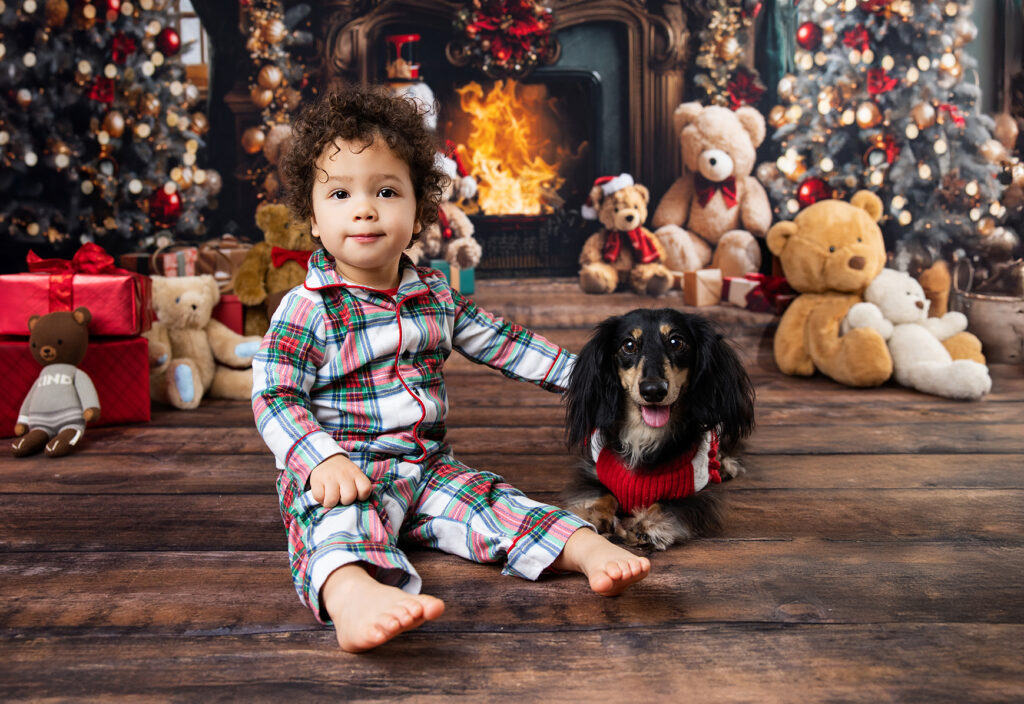Young boy sitting beside a small dog in front of a festive Christmas fireplace with teddy bears