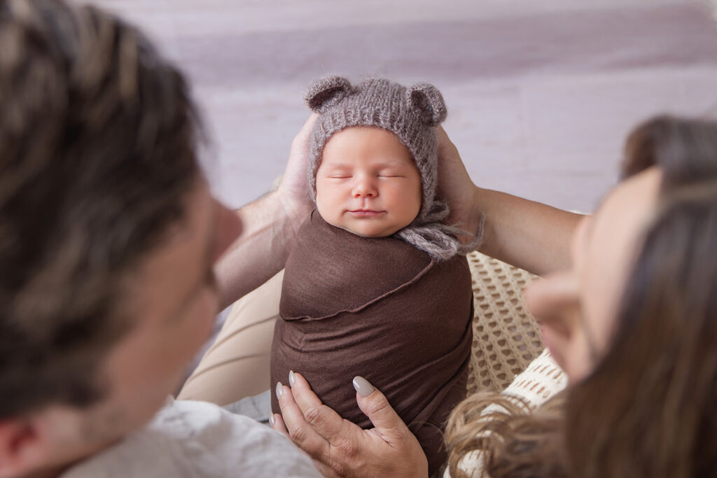 Baby wrapped in a soft brown blanket wearing a knitted bear hat, gently supported by loving hands during a newborn photography session.