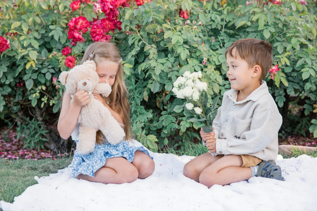 East Bay family photography: Two young siblings playing together in a flower garden, with the girl holding a teddy bear as warm evening sunlight illuminates the scene.