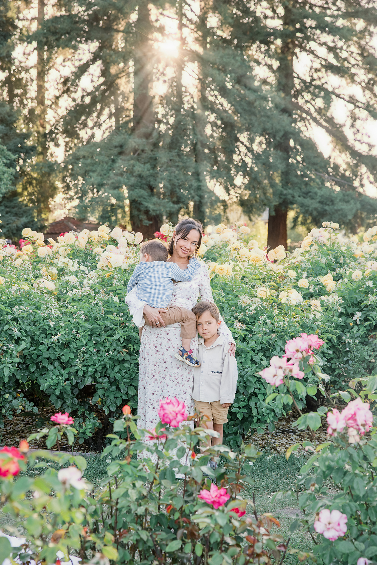 Golden hour family photography: Mother holding her baby while standing beside her young daughter in a blooming garden, surrounded by soft evening light.