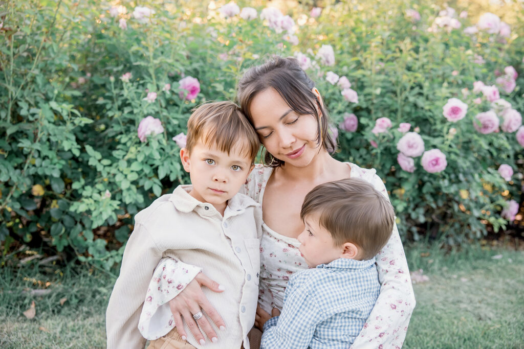 Mother embracing her two children in a blooming garden during golden hour, with soft evening light creating warm family portraits.