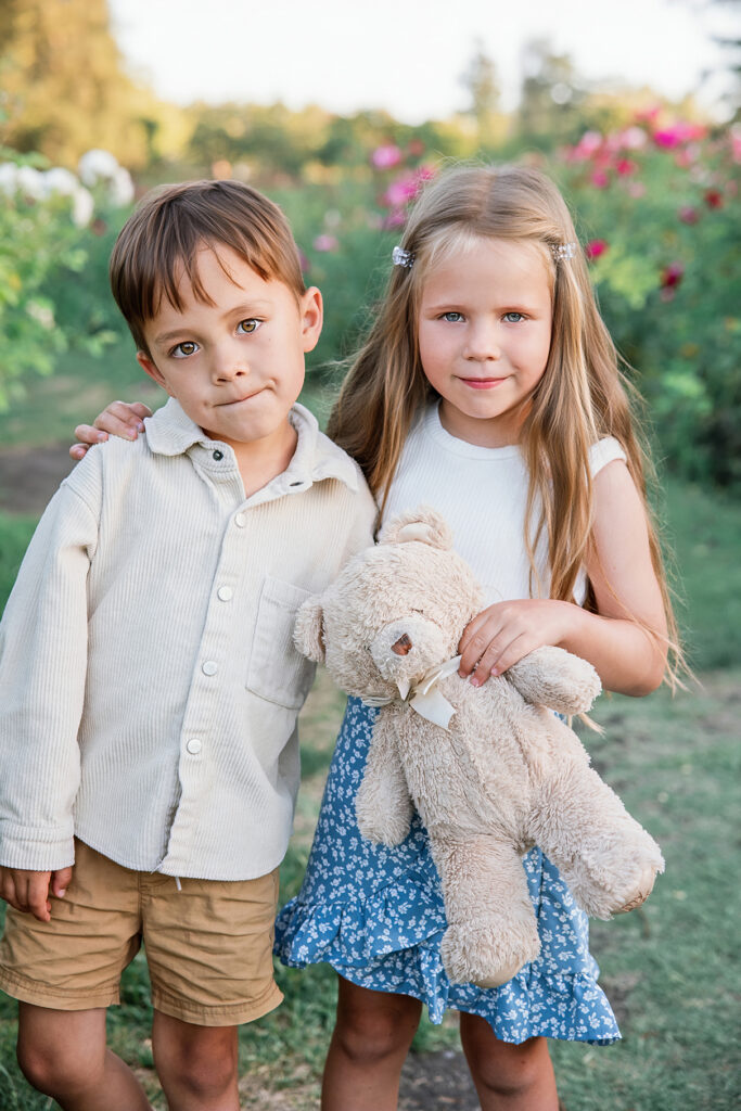 Golden hour family session: Young boy and girl standing together in a garden, with the girl holding a teddy bear and both smiling at the camera.