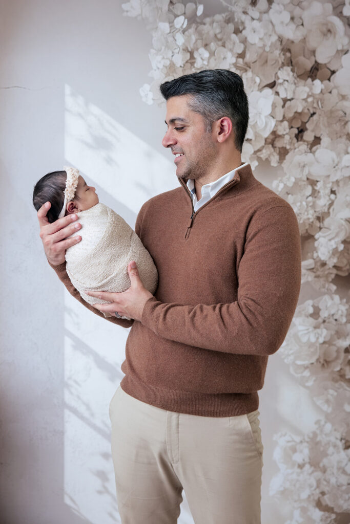 Father smiling while holding his newborn baby girl in a soft, neutral studio setting at FrameIt Bay Area.