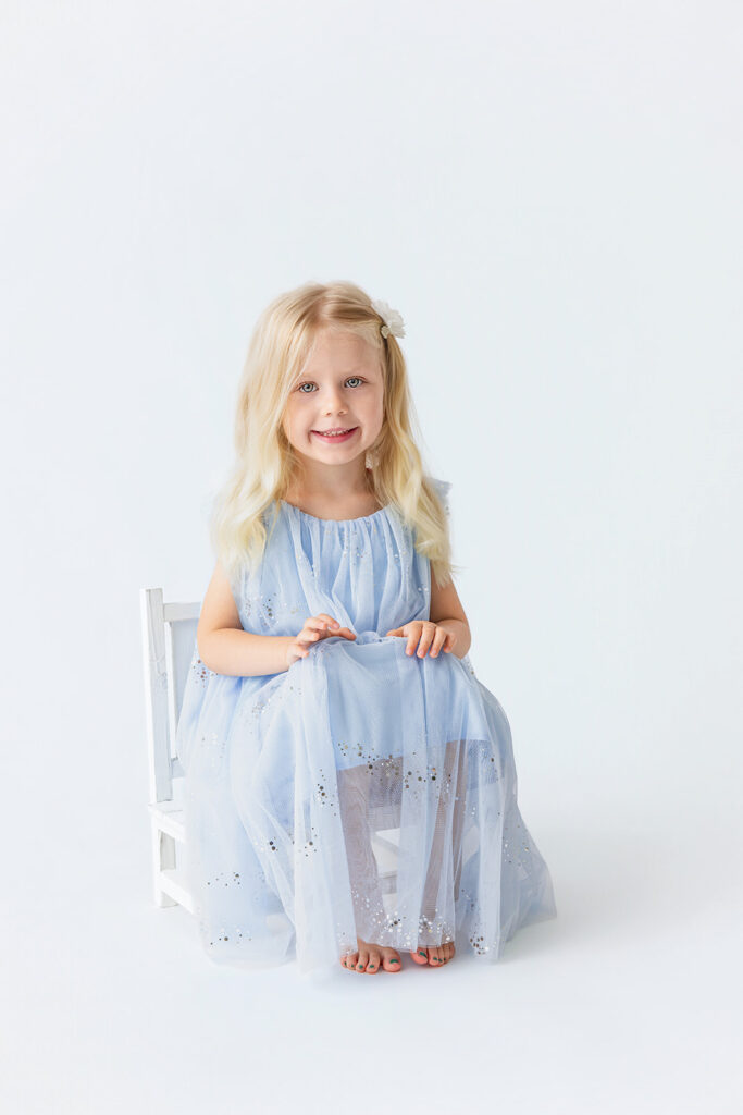 Smiling little girl in a blue dress sitting in a studio, illustrating family photoshoot tips for capturing natural expressions.