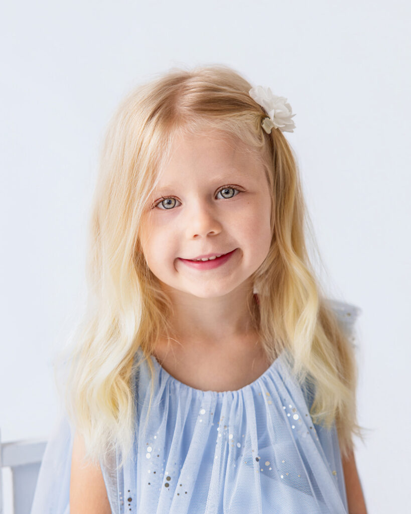 Close-up portrait of a little girl with blonde hair and a white floral hair accessory, wearing a blue dress.