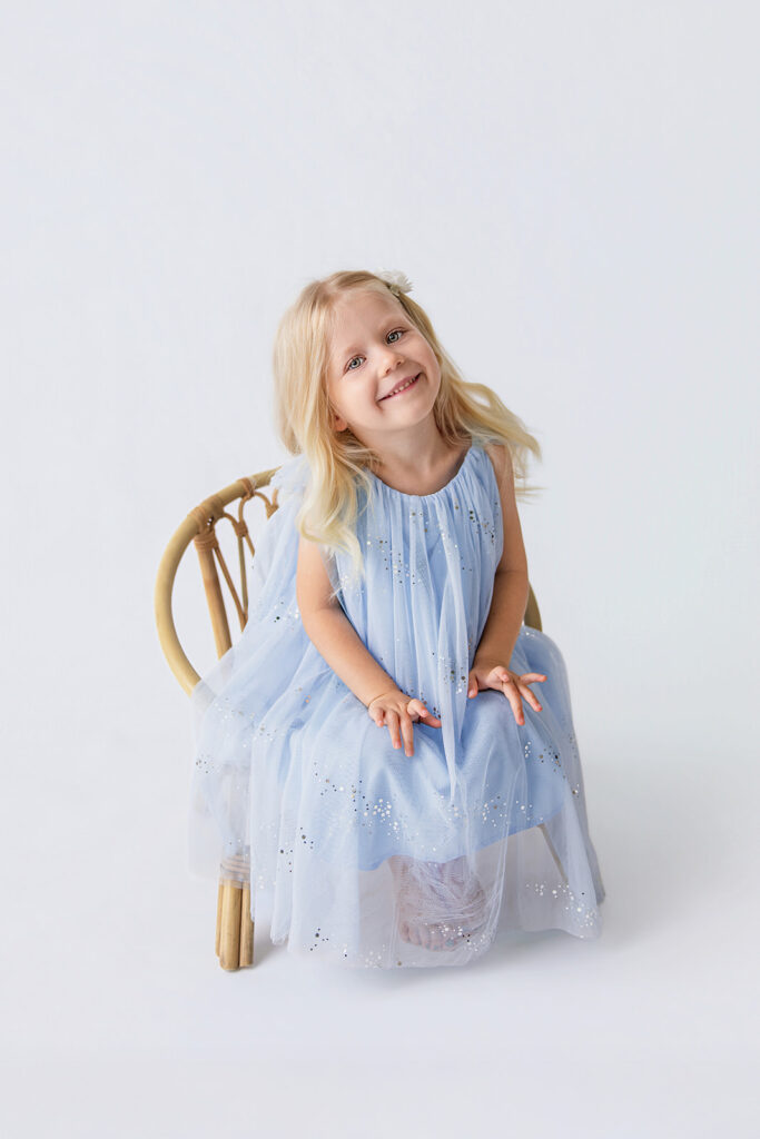 Happy little girl sitting on a wooden chair in a blue dress during a children’s photography session in the Bay Area.