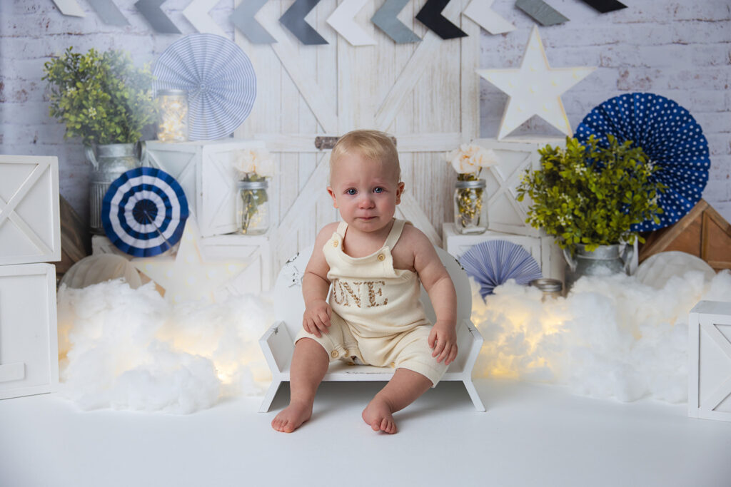 Baby boy sitting and looking toward the camera during a first birthday milestone photo session with blue-and-white decorations.